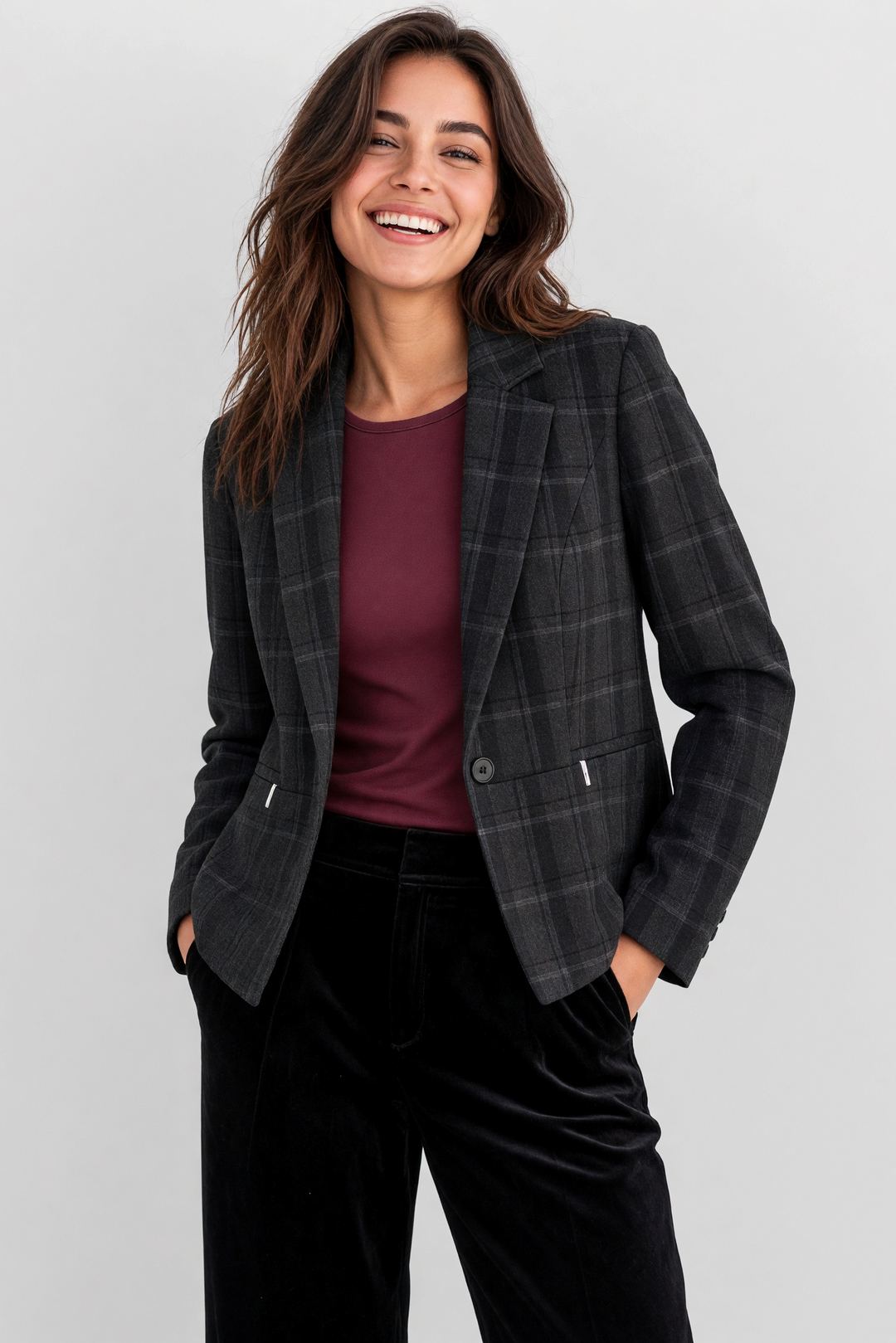 A woman smiling and posing in a dark gray plaid blazer over a burgundy top and black velvet trousers, photographed against a light gray studio background.