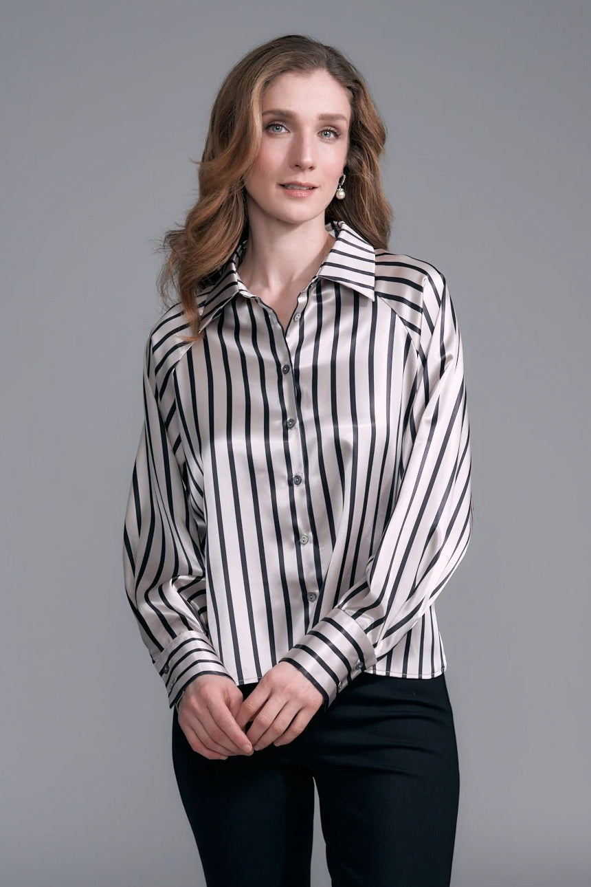 A woman modeling a black and white vertically striped satin button-down shirt with black trousers, standing against a neutral gray studio background.
