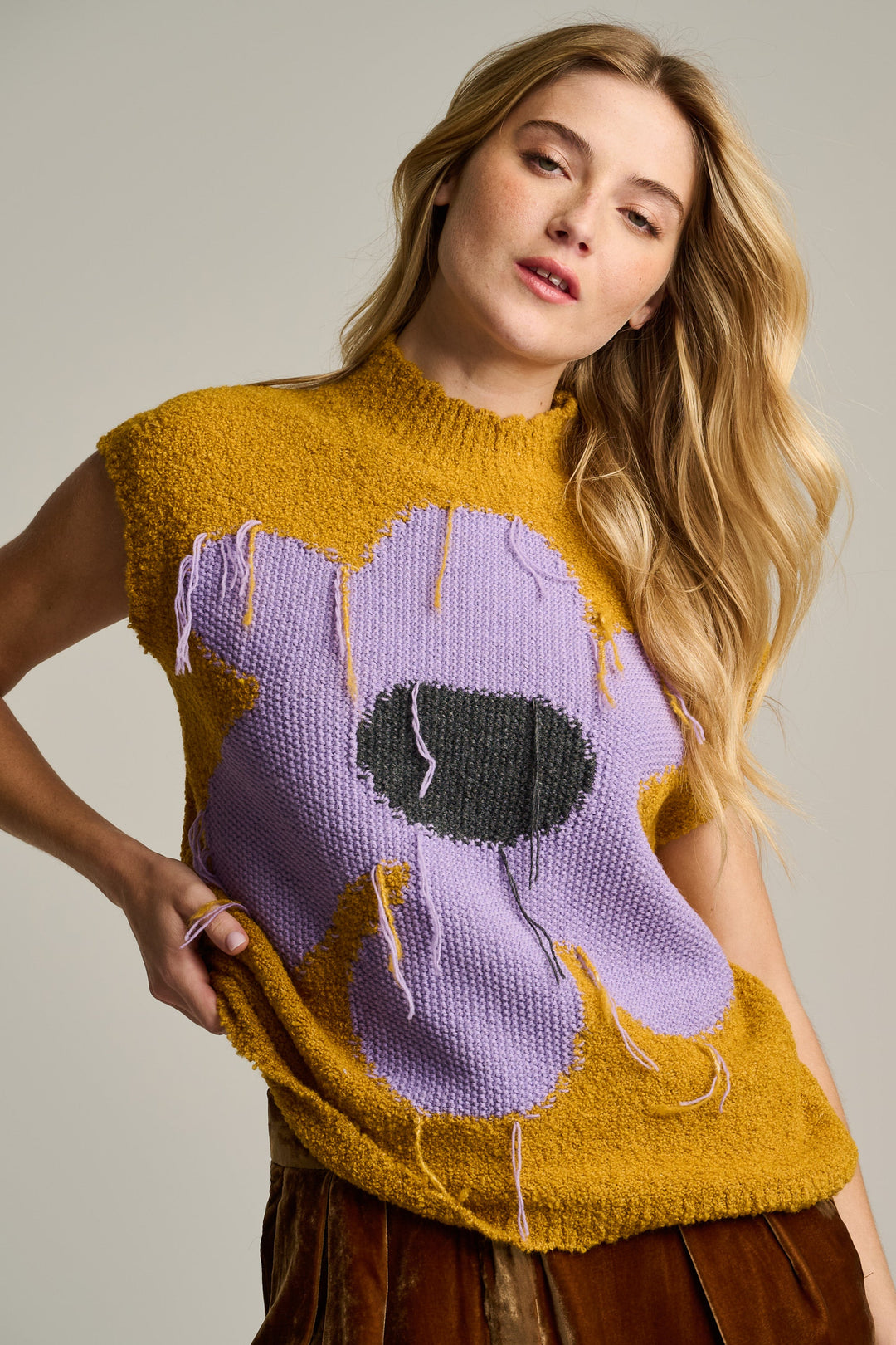 A woman modeling a sleeveless mustard yellow knit sweater with a large lavender and dark gray abstract graphic on the front, paired with brown bottoms, against a plain light gray studio background.