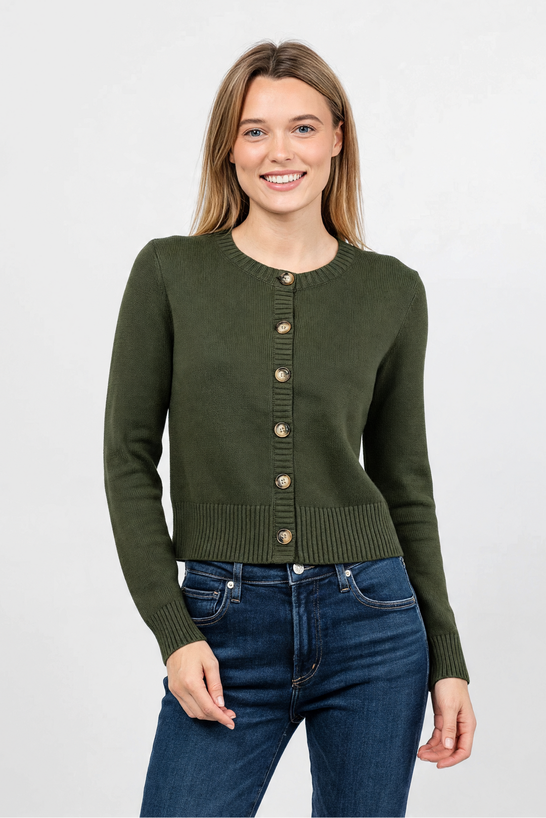 A woman modeling a dark olive green cropped cardigan sweater with gold-tone buttons, paired with dark blue jeans, against a plain light gray studio background.